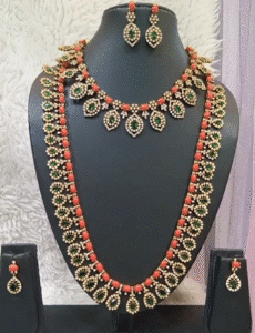 Coral Beaded Bridal Jewellery  with red coral beads, green Kundan stones, gold detailing, including two layered necklaces, a pair of dangler earrings, and stud earrings displayed on a black stand